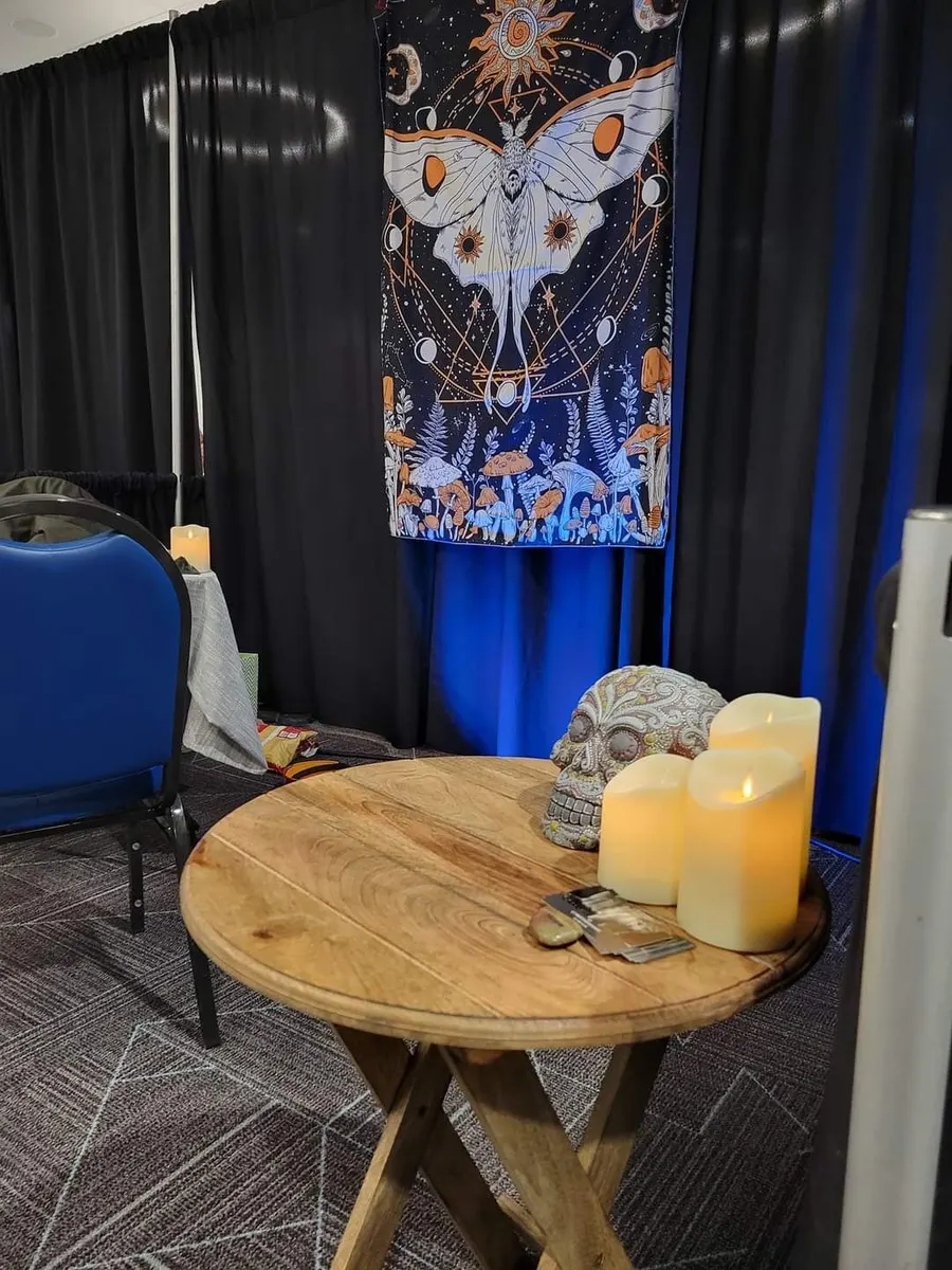 Crystal’s tarot table at the Oregon Ghost Conference: a round wooden table with candles and a sugar skull under a luna-moth tapestry.